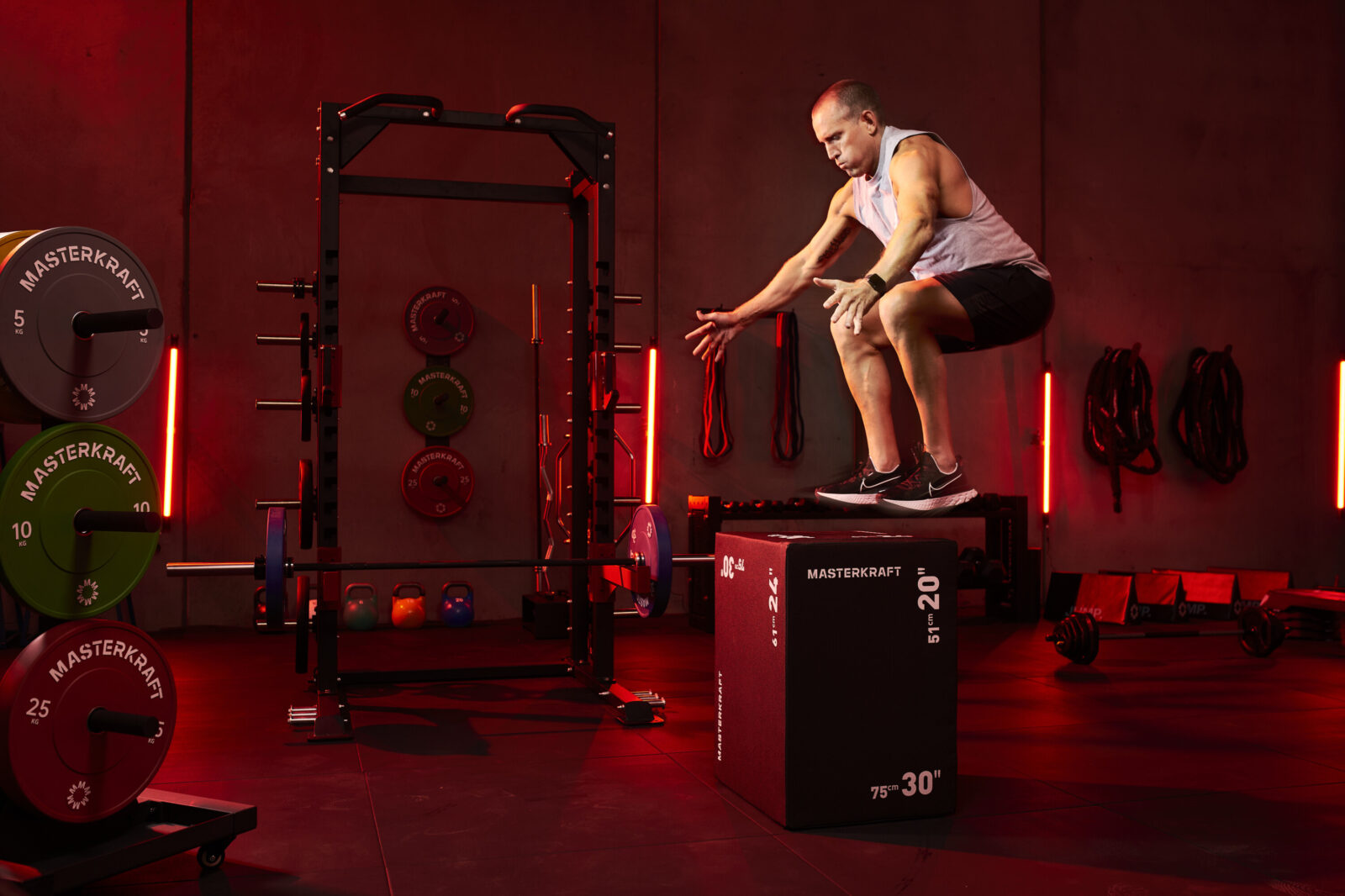 Box jump onto a MasterKraft plyo box beside a half rack, bumper plates and kettlebells