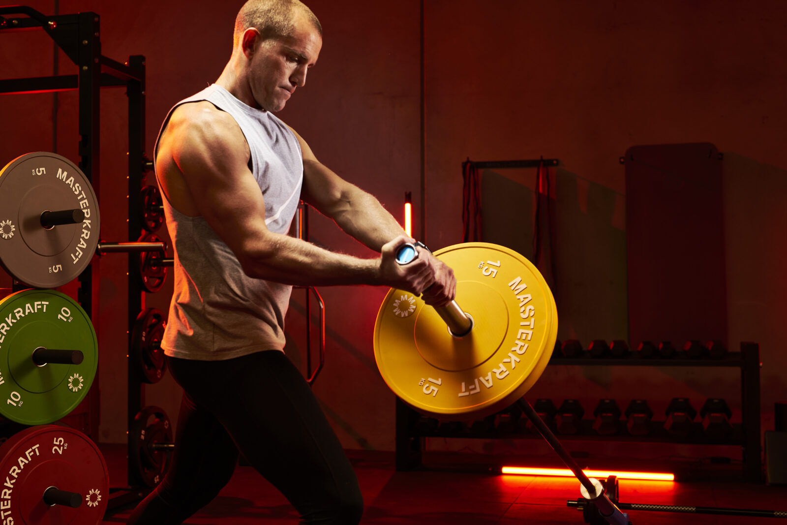 Athlete swinging a MasterKraft barbell with 15 kg bumper plates beside a loaded rack